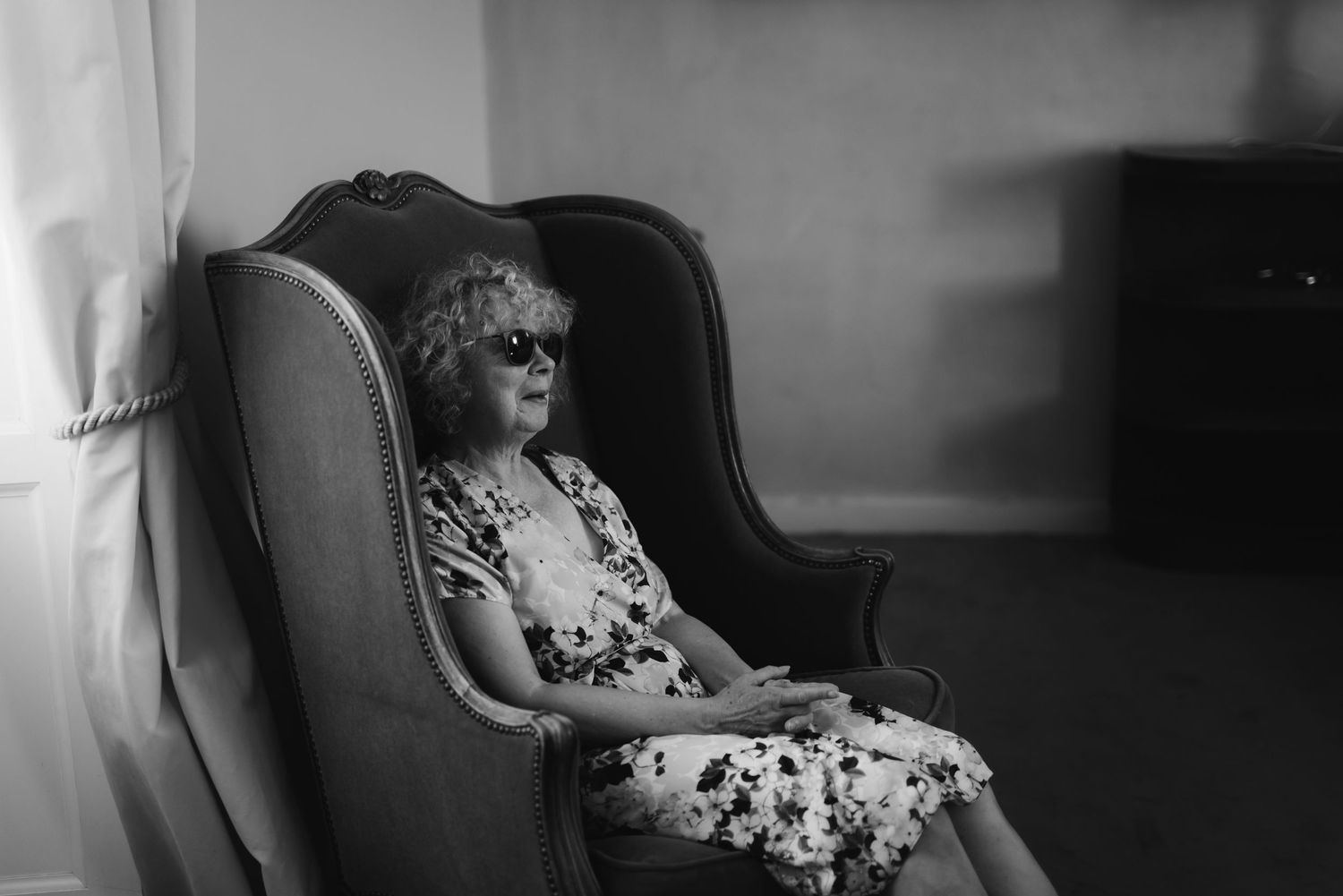 Black and white portrait of someone seated in an elegant armchair wearing a floral dress.