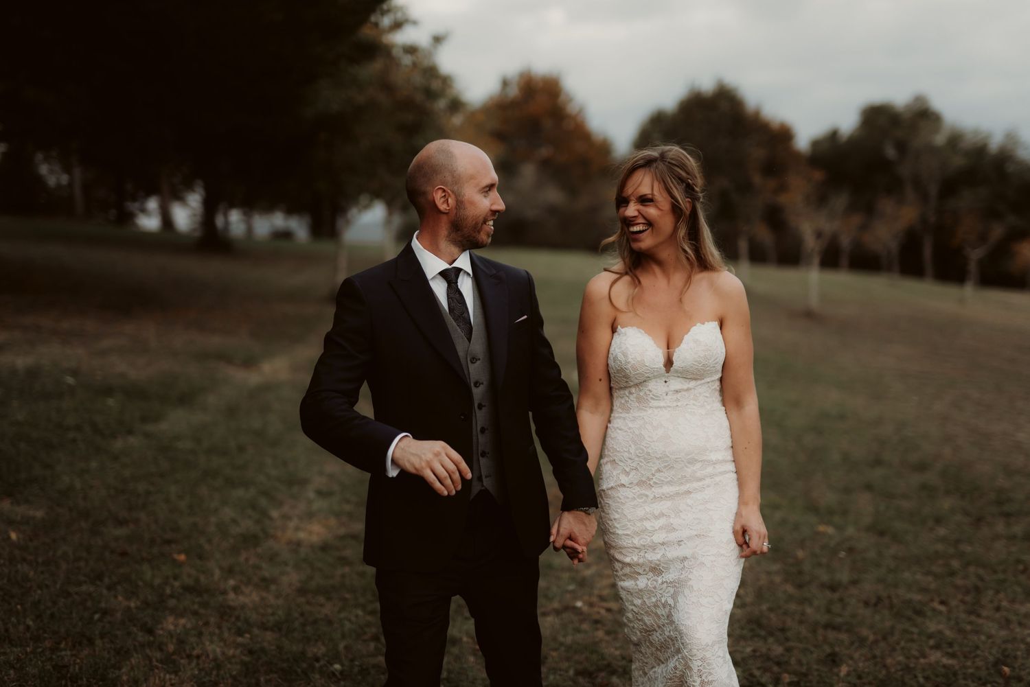 A couple laughs together in formal attire on a grassy autumn hillside.