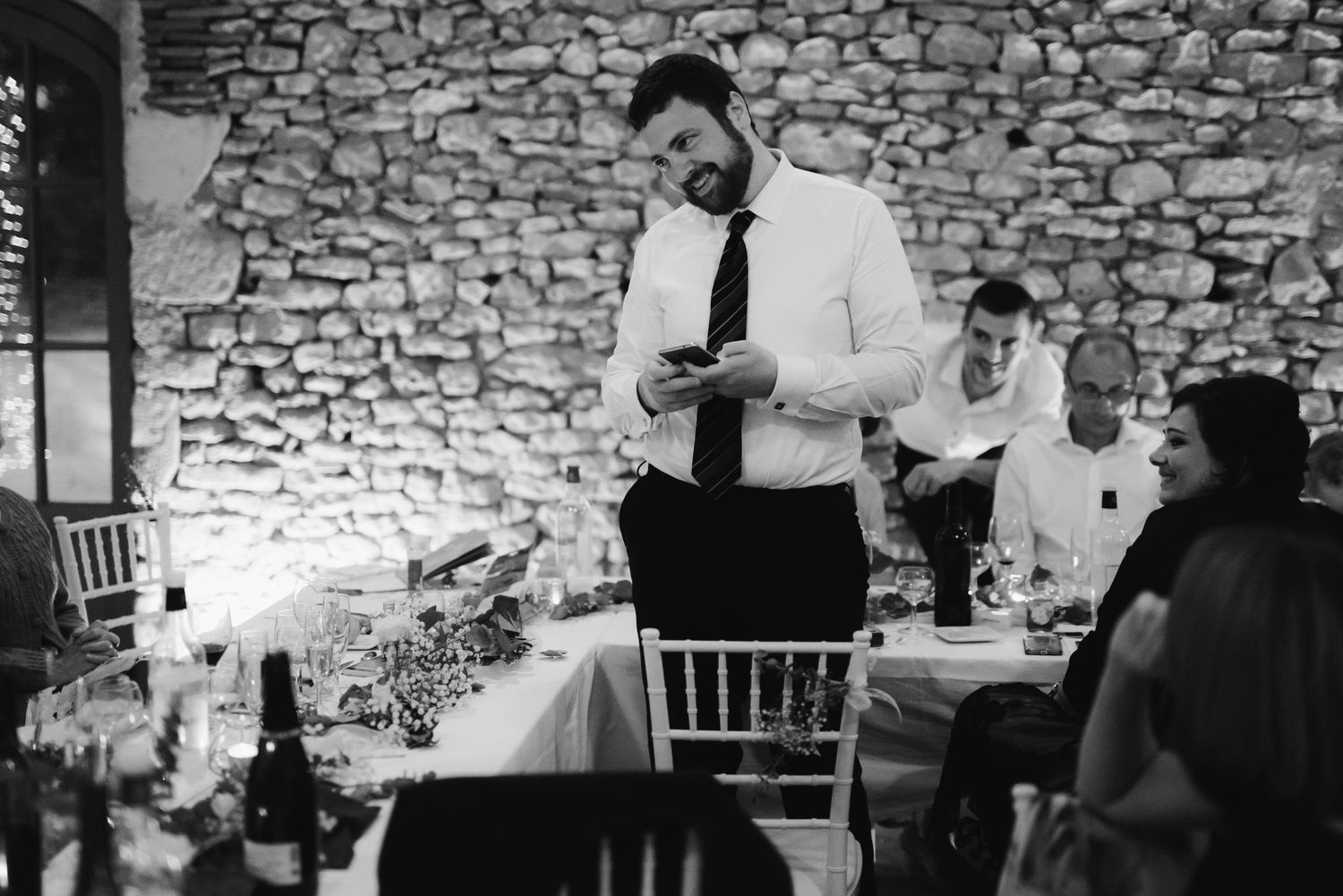A reception speech is delivered in black and white at an elegant stone-walled wedding venue.