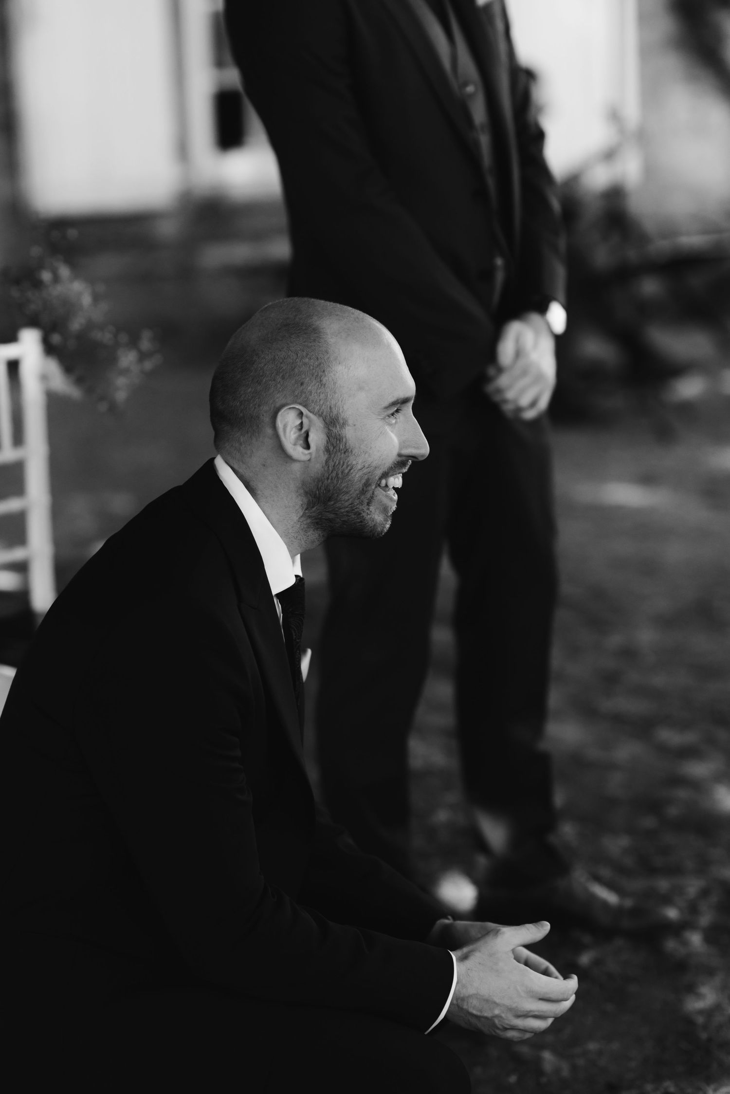A man in a black suit smiles during an outdoor wedding ceremony.