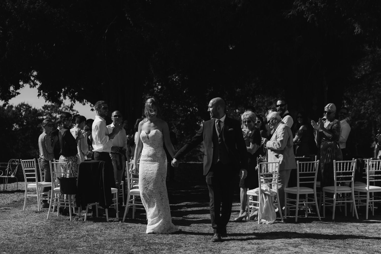 A black and white capture of a wedding ceremony with guests seated in white chairs under trees.