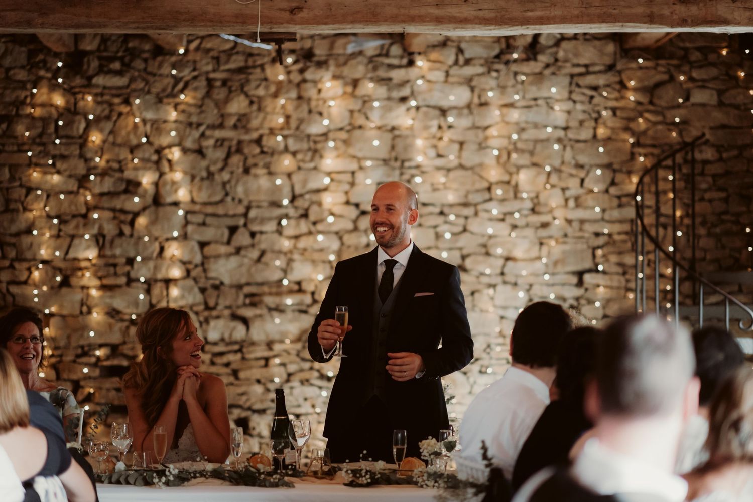 Wedding reception speech in a rustic stone-walled room illuminated by fairy lights.