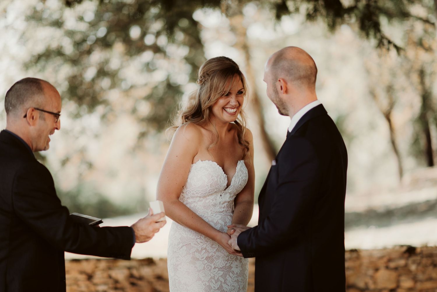 A couple smiles during their wedding ceremony as they exchange vows under a shaded outdoor canopy.