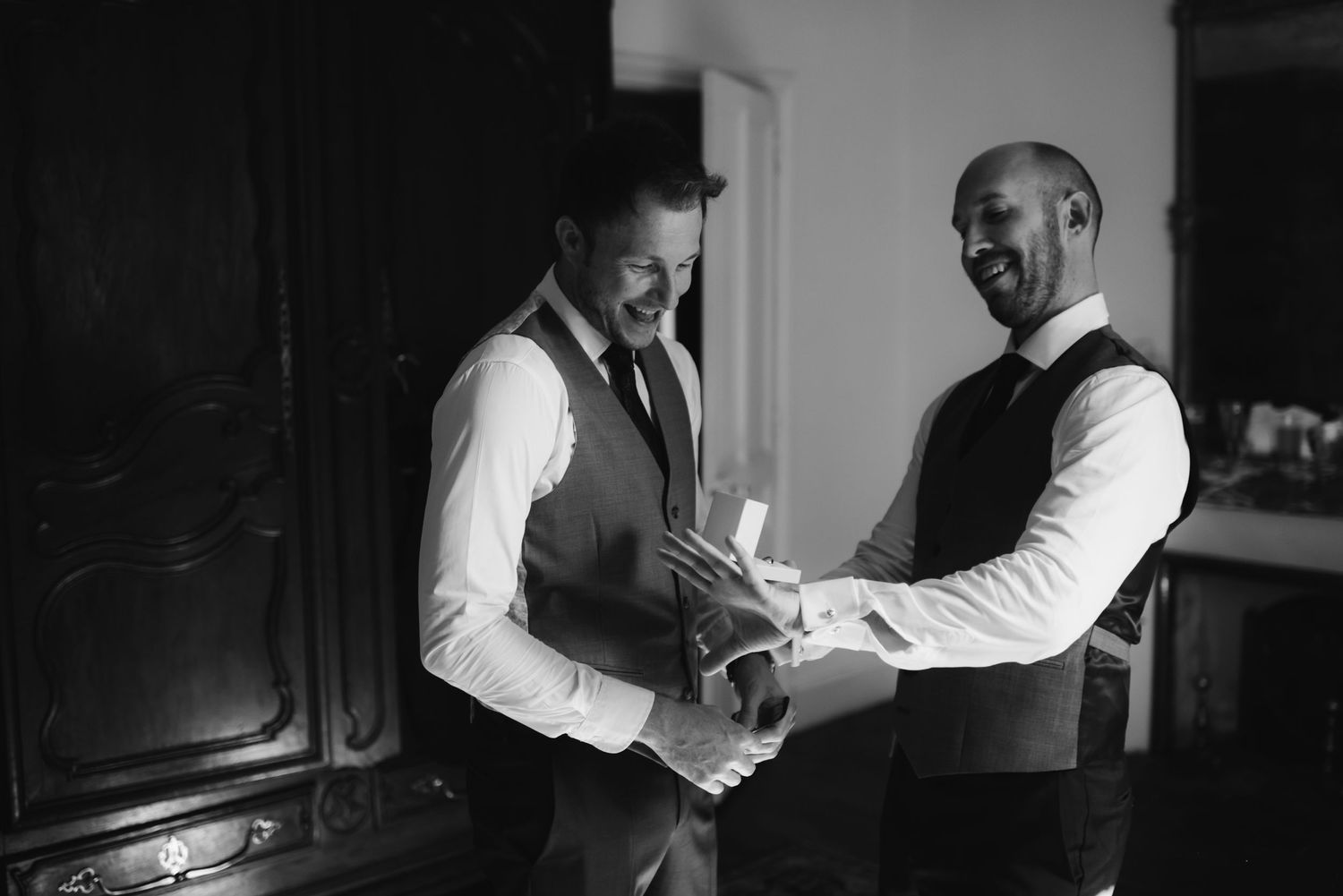 Black and white image of two men in vests helping each other with formal preparations.