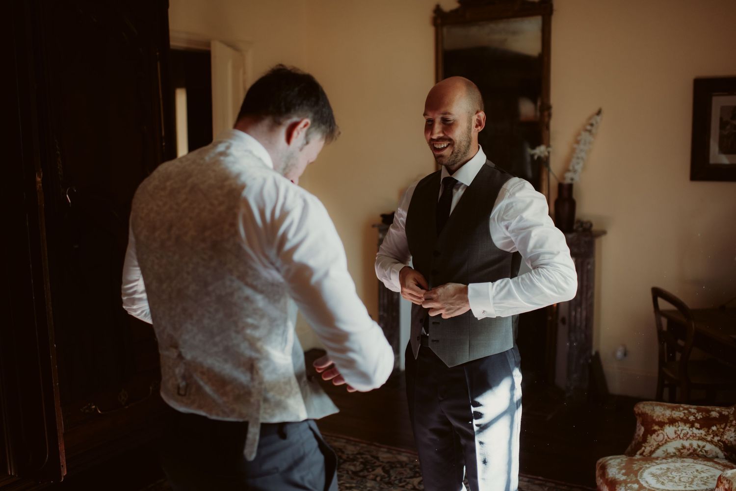 Two men in formal wear laughing while getting dressed in a traditional room.