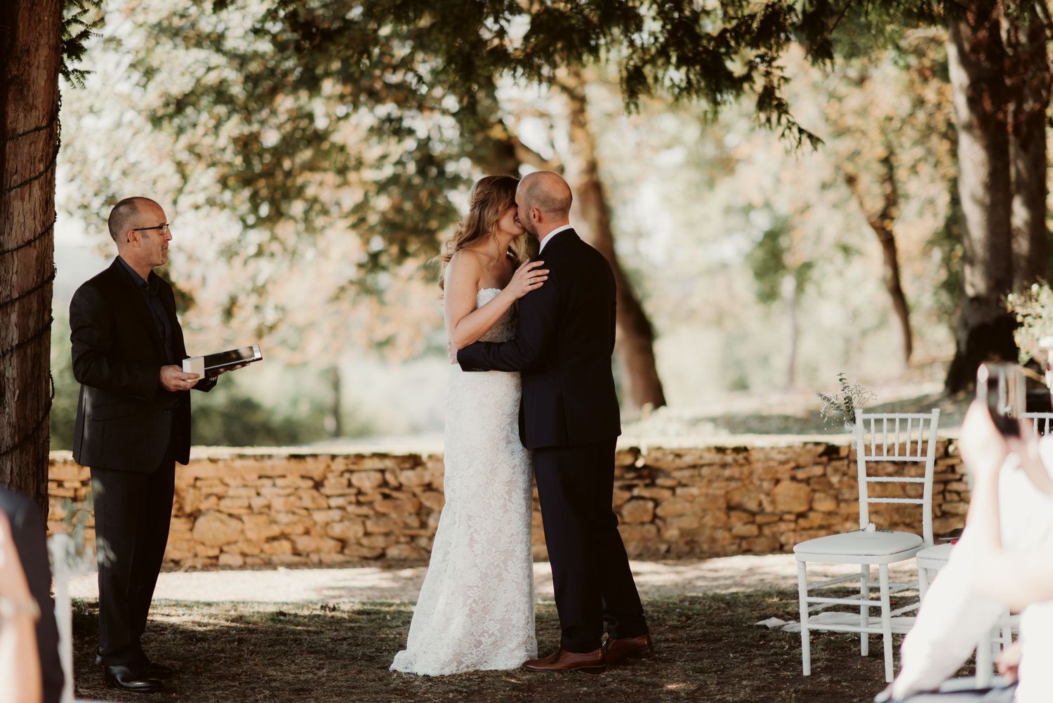 An intimate outdoor wedding ceremony unfolds under tall trees with white chairs and a stone wall backdrop.