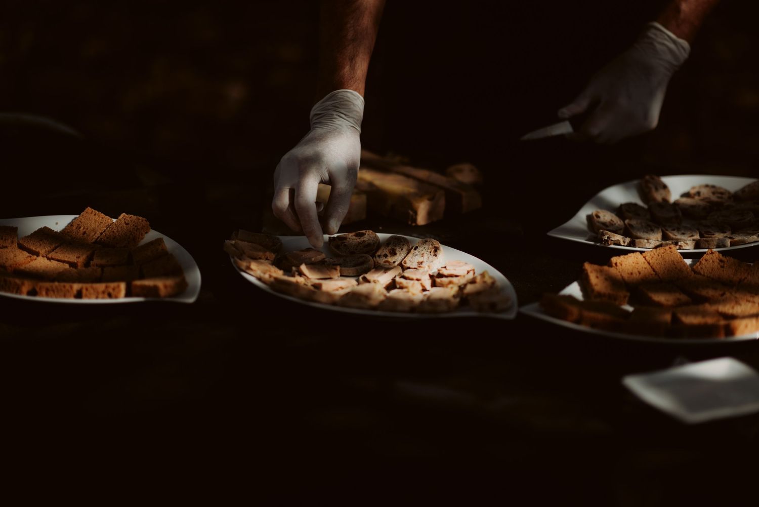 Gloved hands prepare food plates in dramatic lighting at a catered event.