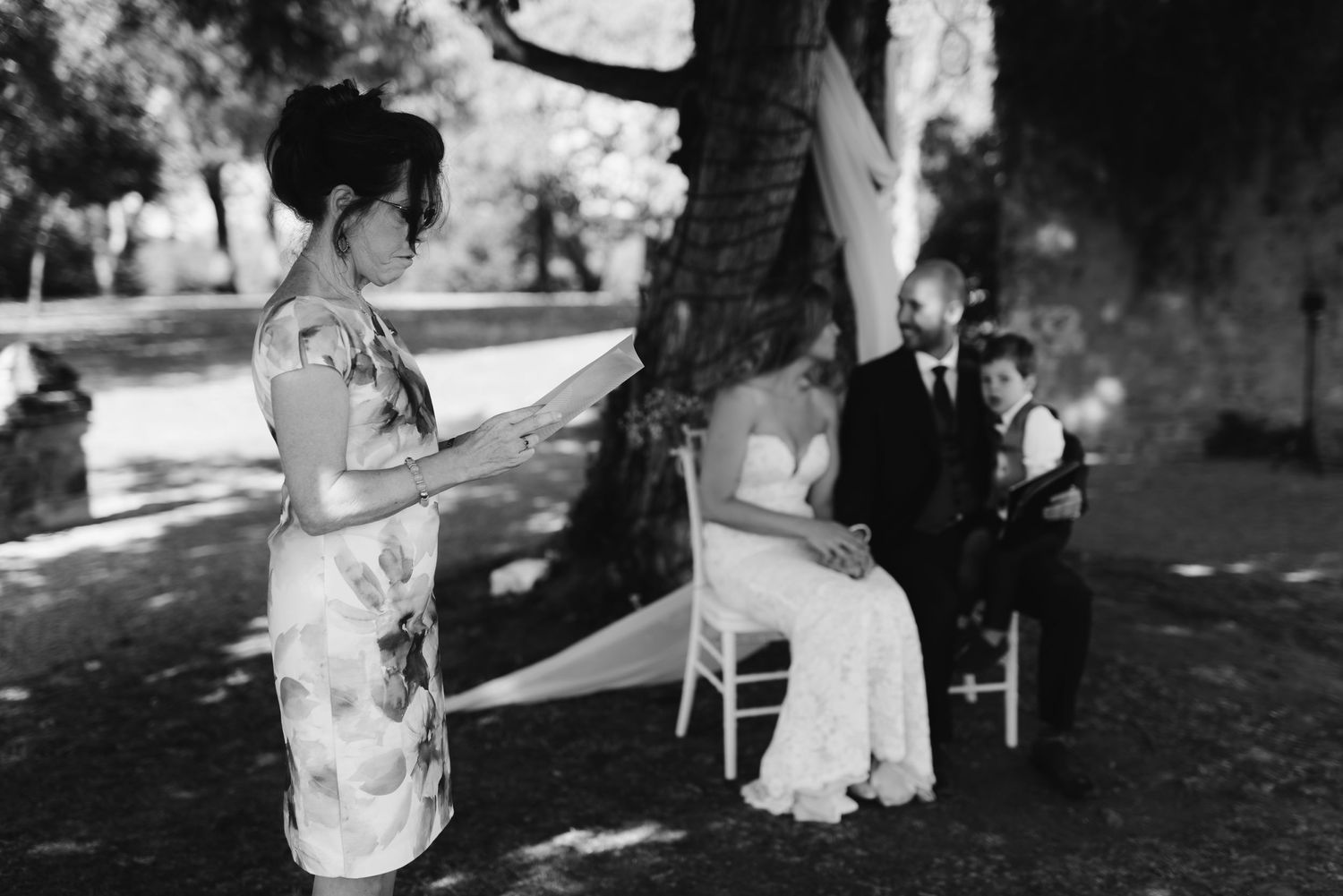 A black and white wedding ceremony photo under a tree with elegant attire and traditional decorations.