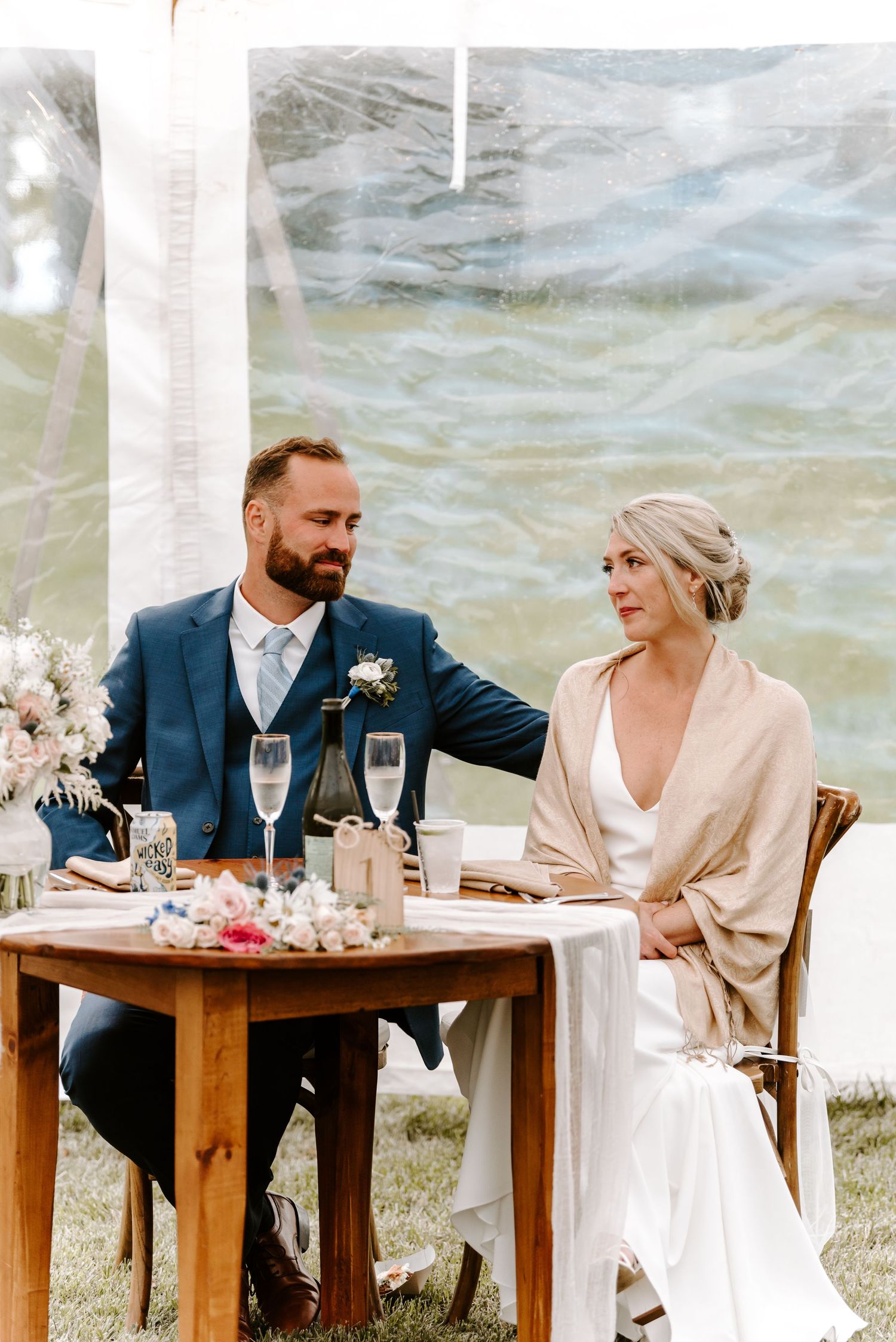 Wedding couple shares an intimate moment at their reception table under an outdoor tent by the water.