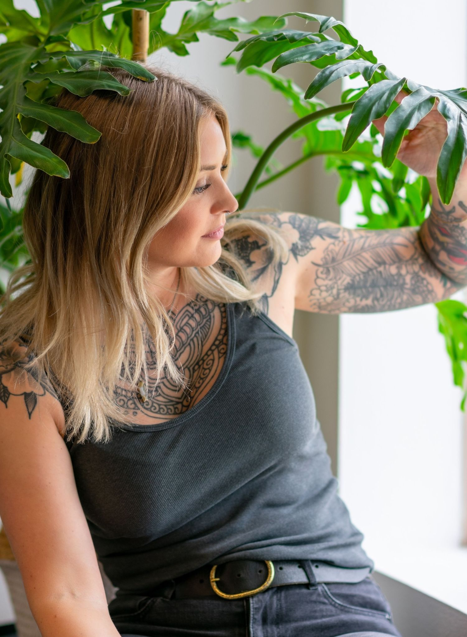 Beautiful woman sitting next to a window lifting up a branch of a plant to look out the window.