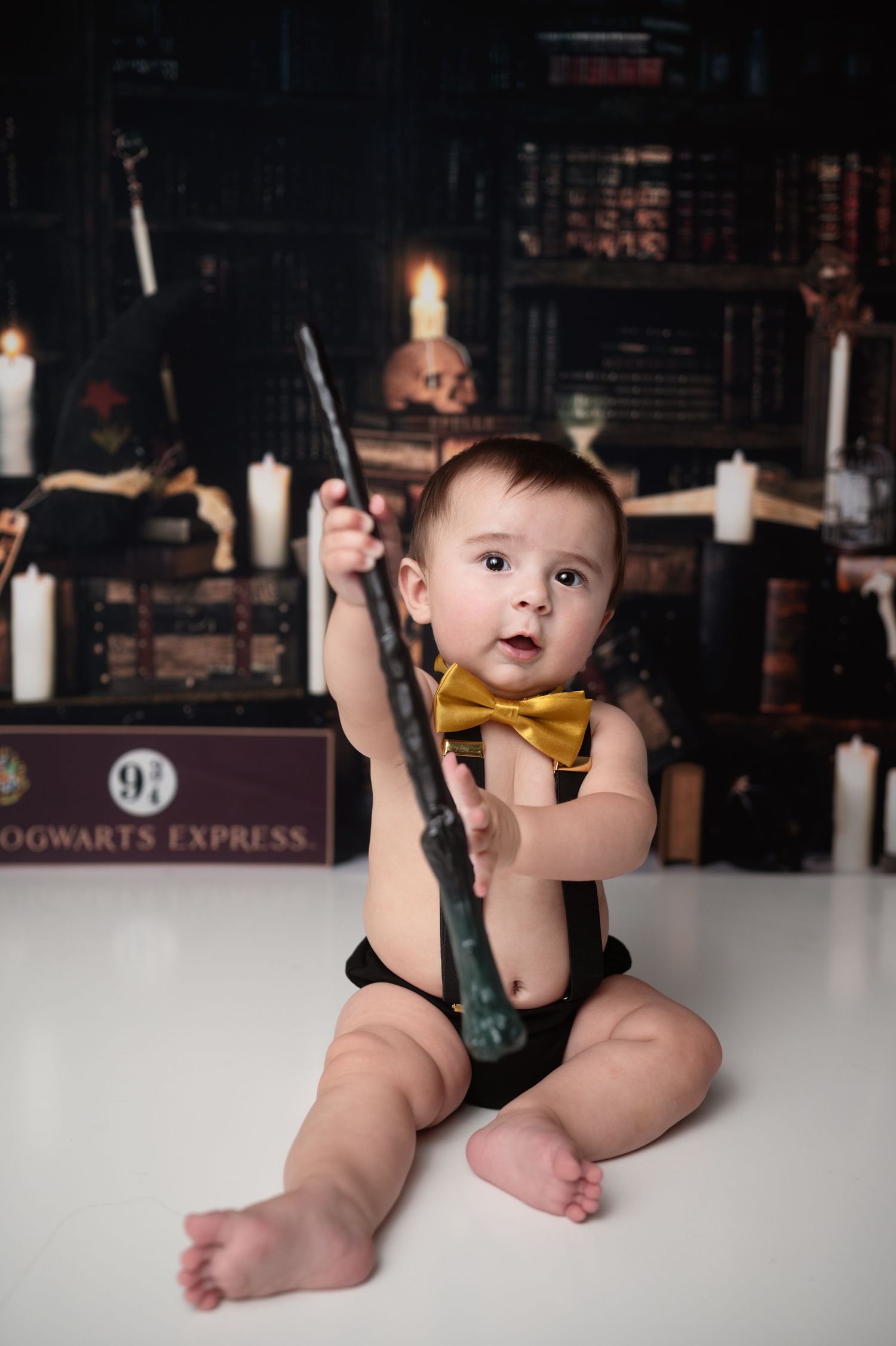 A Harry Potter themed baby photo with wand and bow tie against a magical library backdrop.