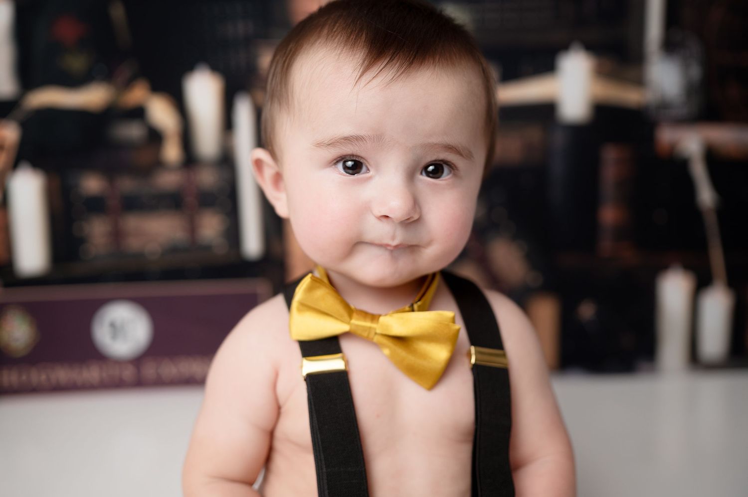Baby wearing black suspenders and yellow bow tie smiles at camera with books in background.