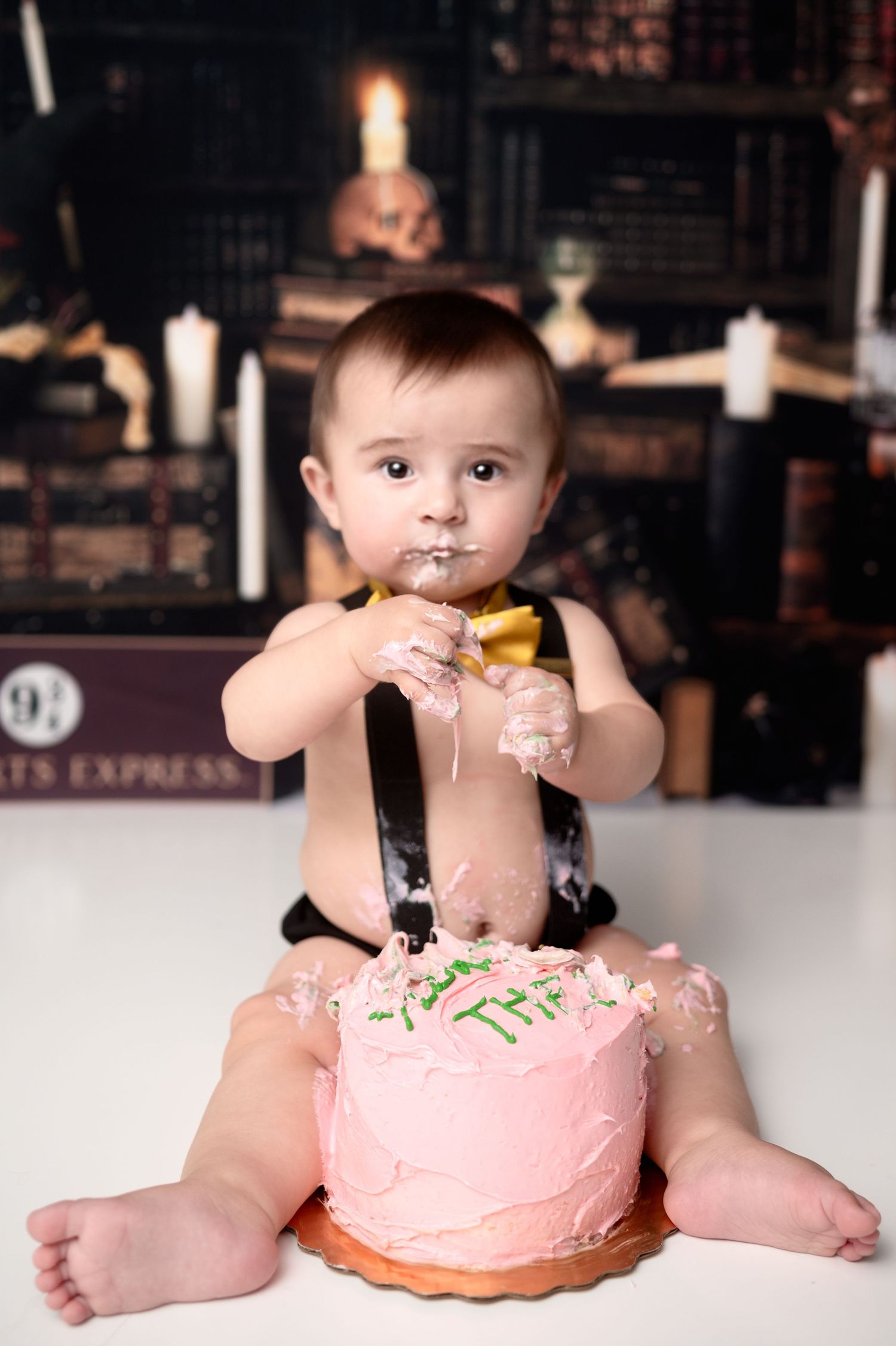 Baby in suspenders enjoying birthday cake smash session with pink frosted cake.