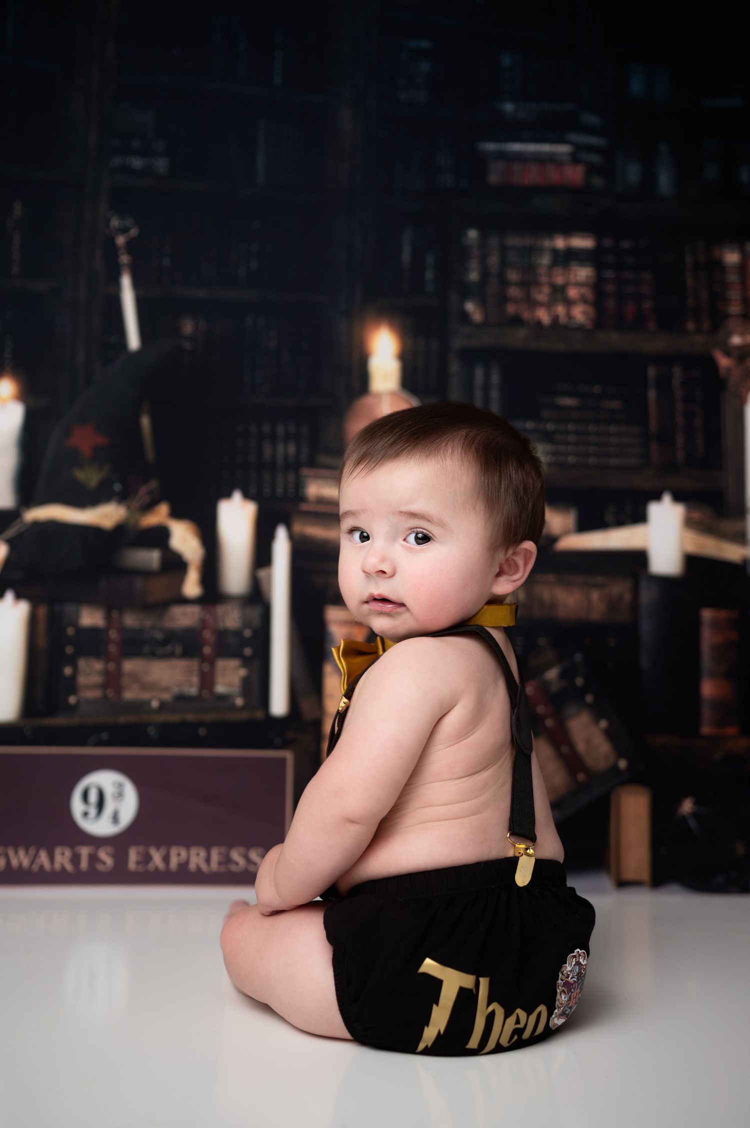 Baby in black diaper cover sits against Harry Potter themed backdrop with candles.