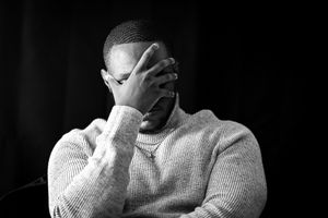 A person in a light sweater covers their face with their hand against a dark background.