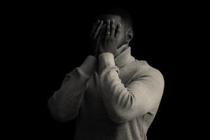 A person in a light sweater covers their face with both hands in dramatic lighting.