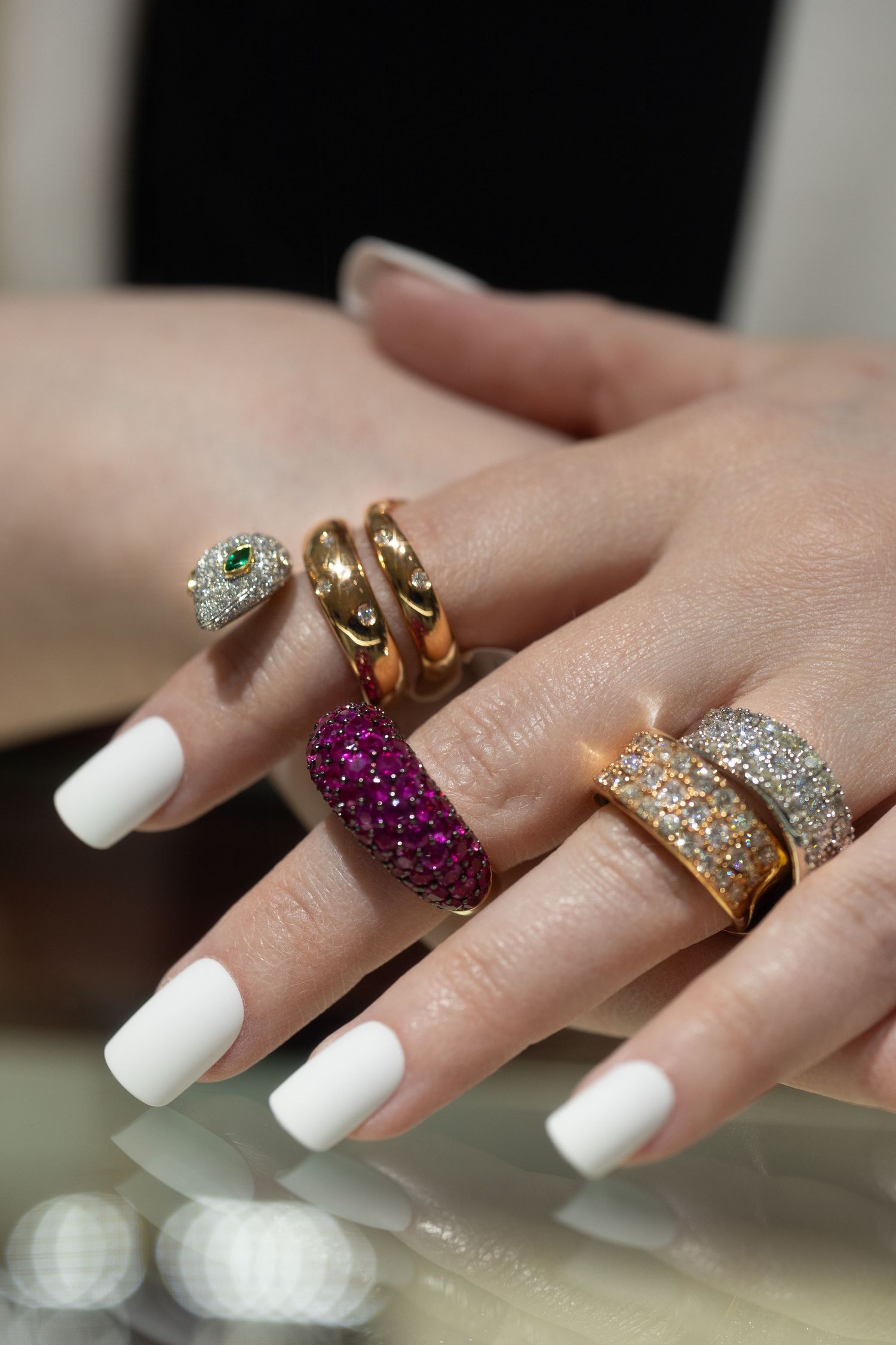 Hand displaying multiple luxurious rings with diamonds and colorful gemstones on white manicured nails.