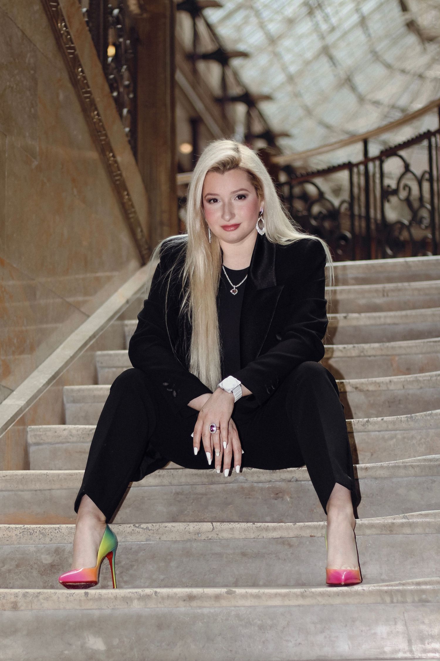 Person in black suit sitting on marble stairs wearing colorful designer heels.