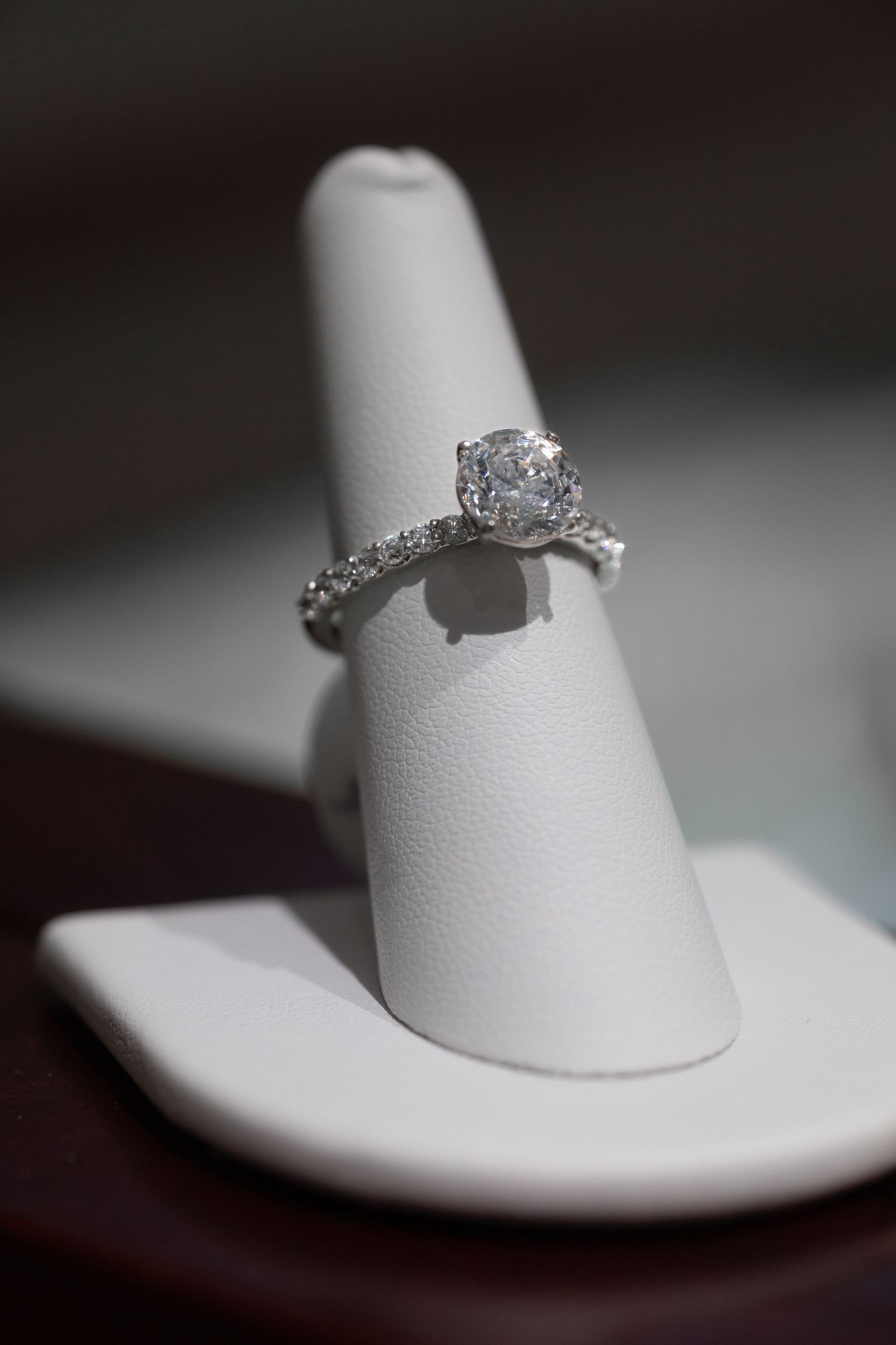 Classic solitaire diamond engagement ring with pave band on white display stand.