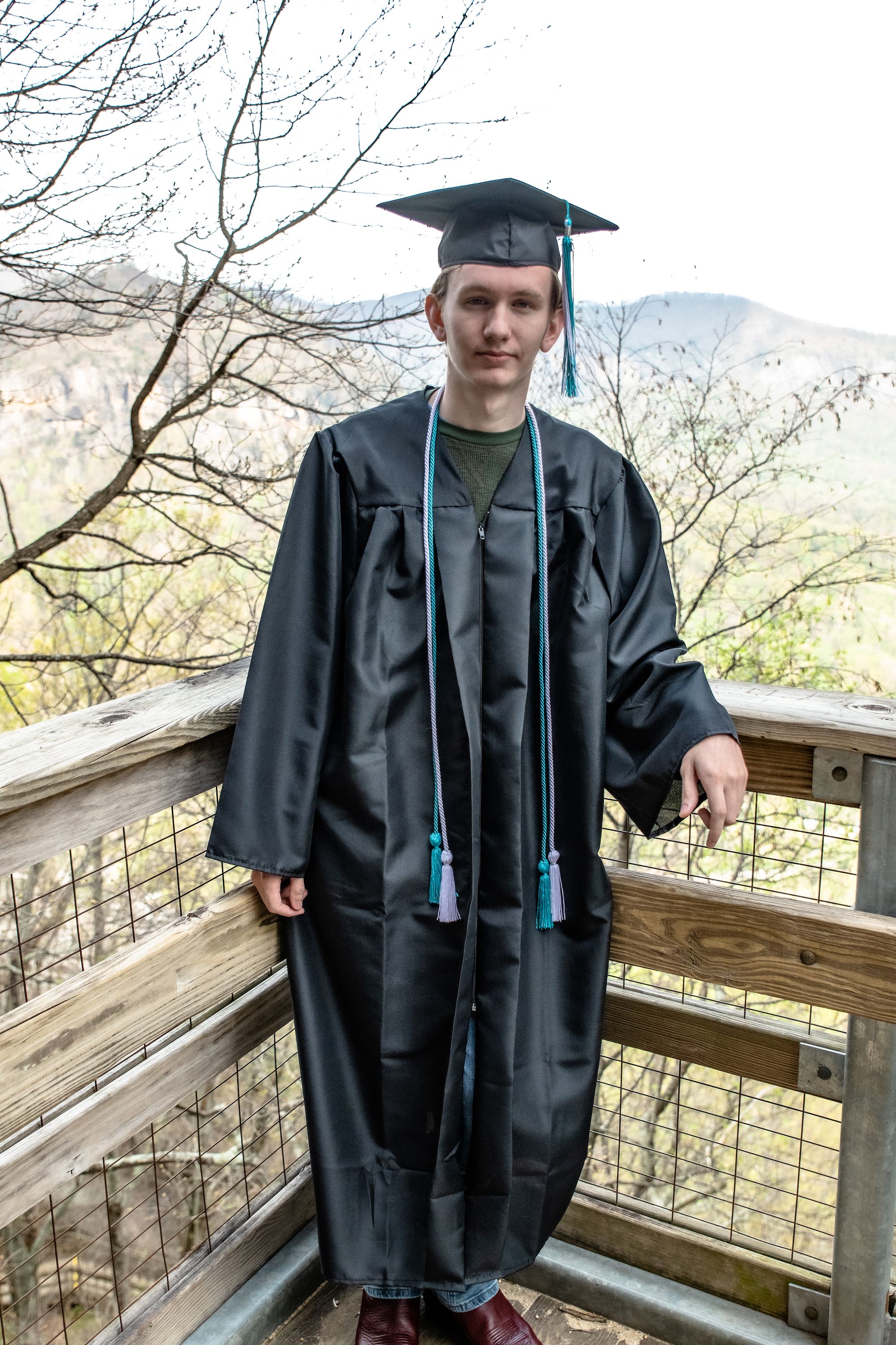 Graduate in traditional black cap and gown standing on wooden deck with mountain view.