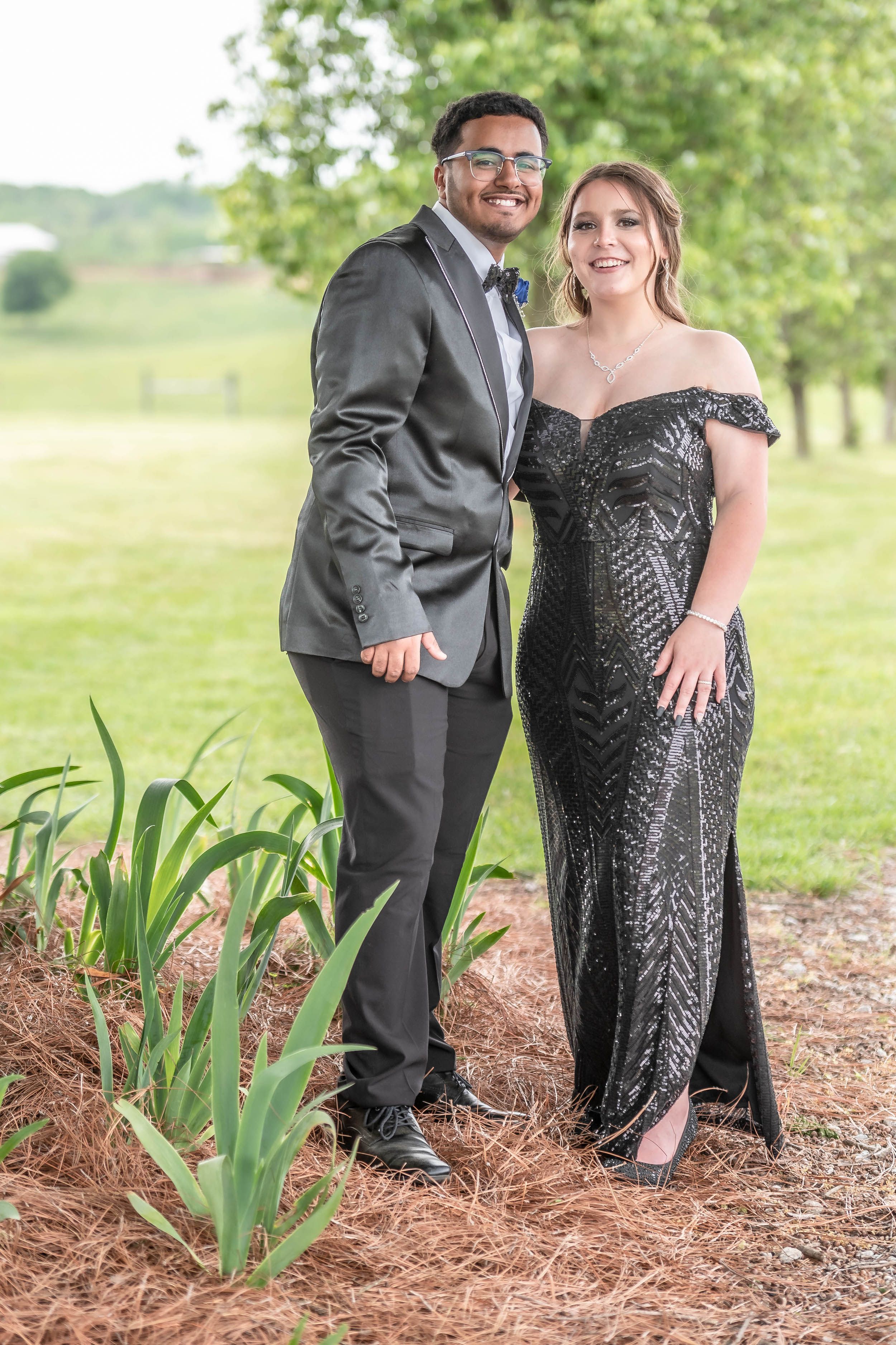 Couple in formal attire poses outdoors, wearing gray suit and black sparkly gown.
