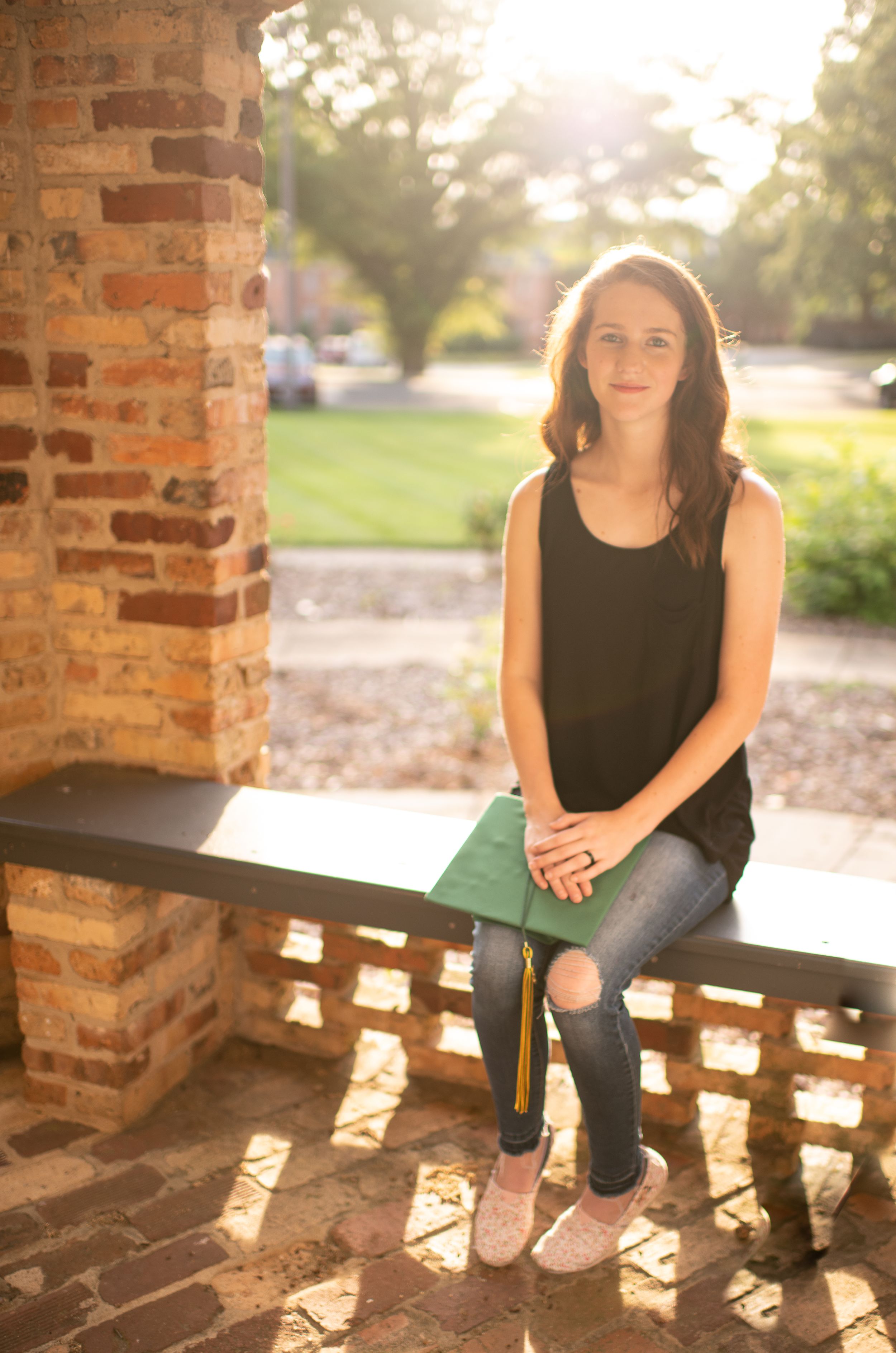 Graduate sitting on brick ledge in casual attire holding graduation cap.
