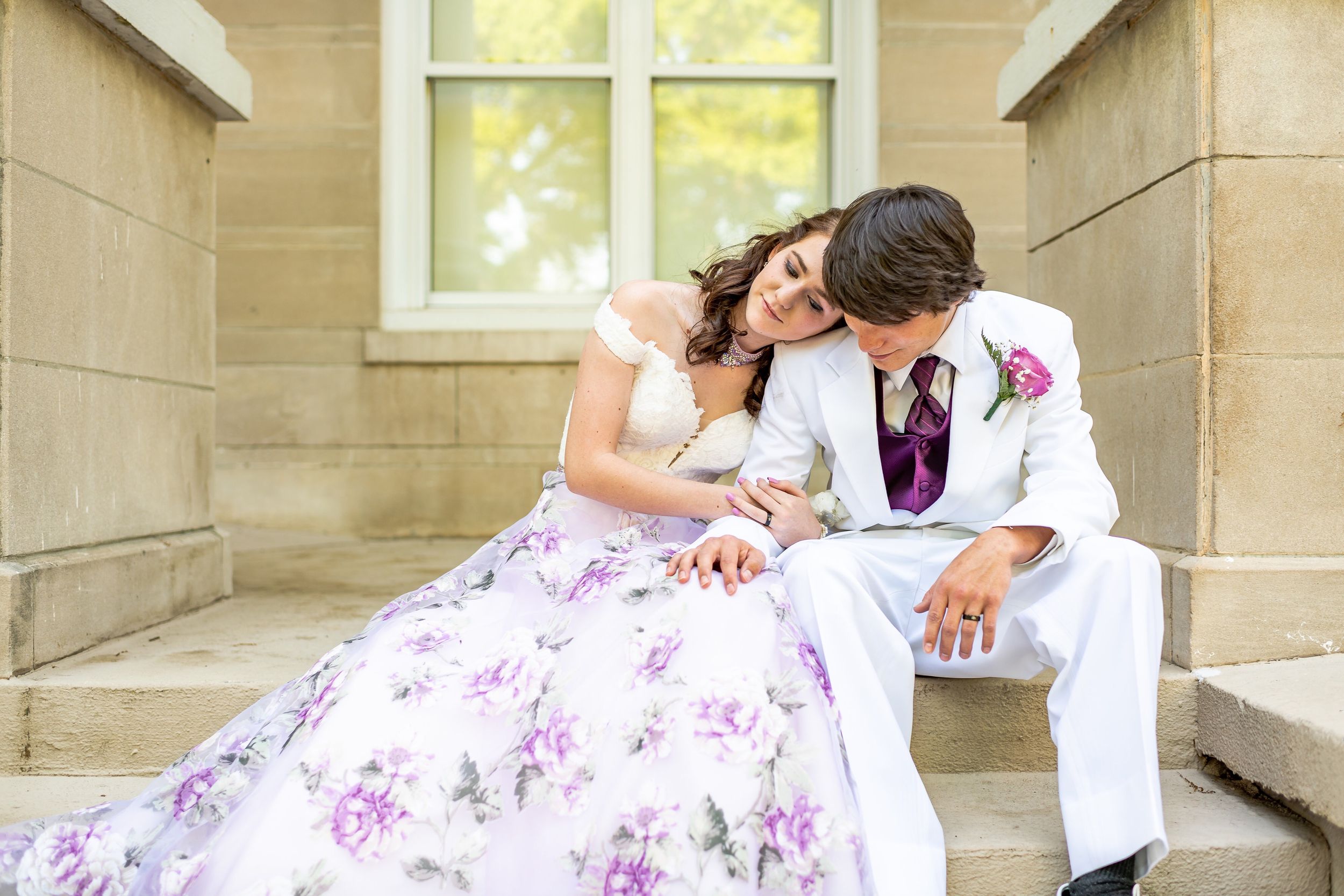 Couple in formal attire resting on stone steps with floral dress details.