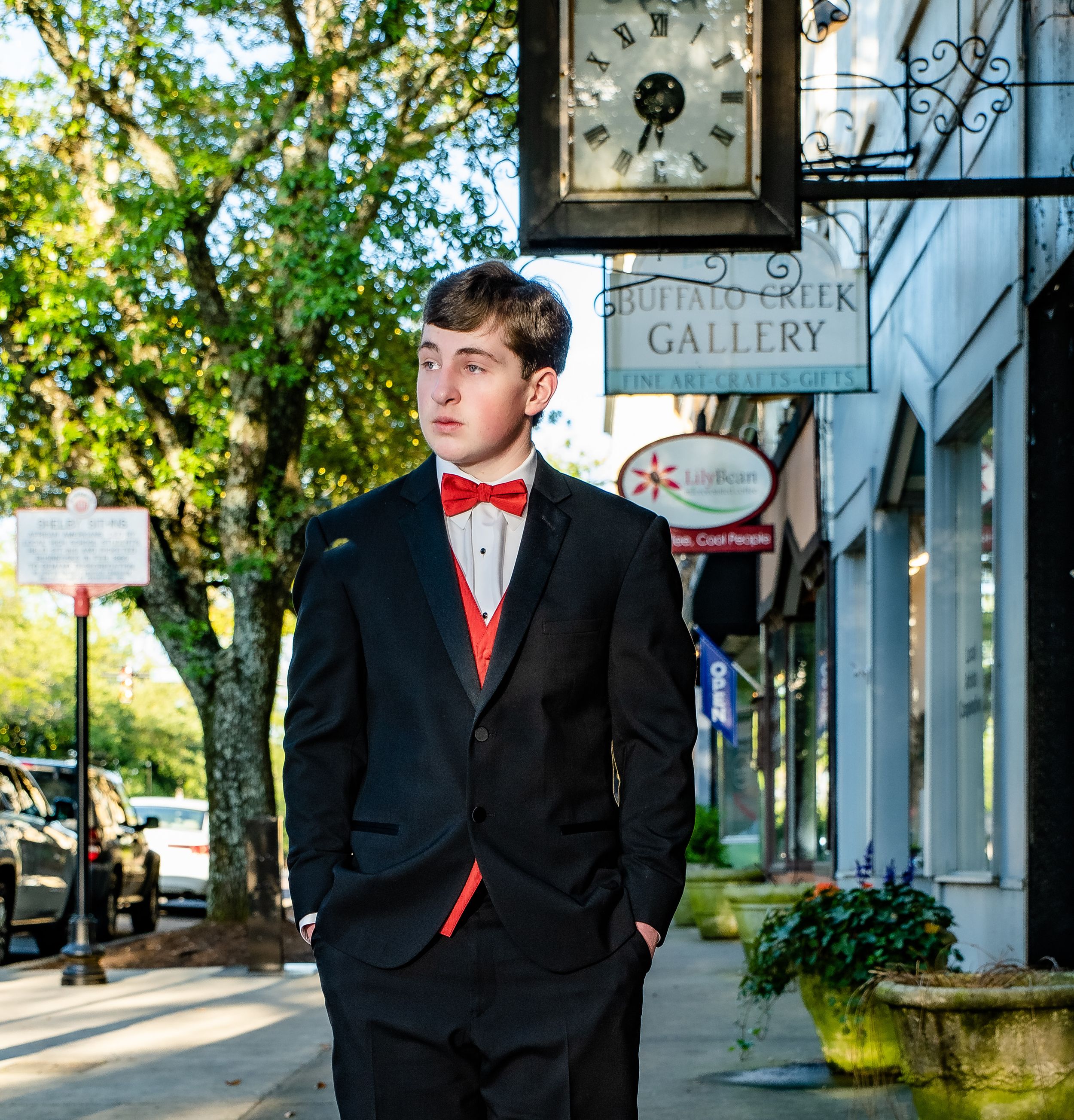 Formal portrait of young man in black tuxedo and red bowtie standing outside gallery storefront.