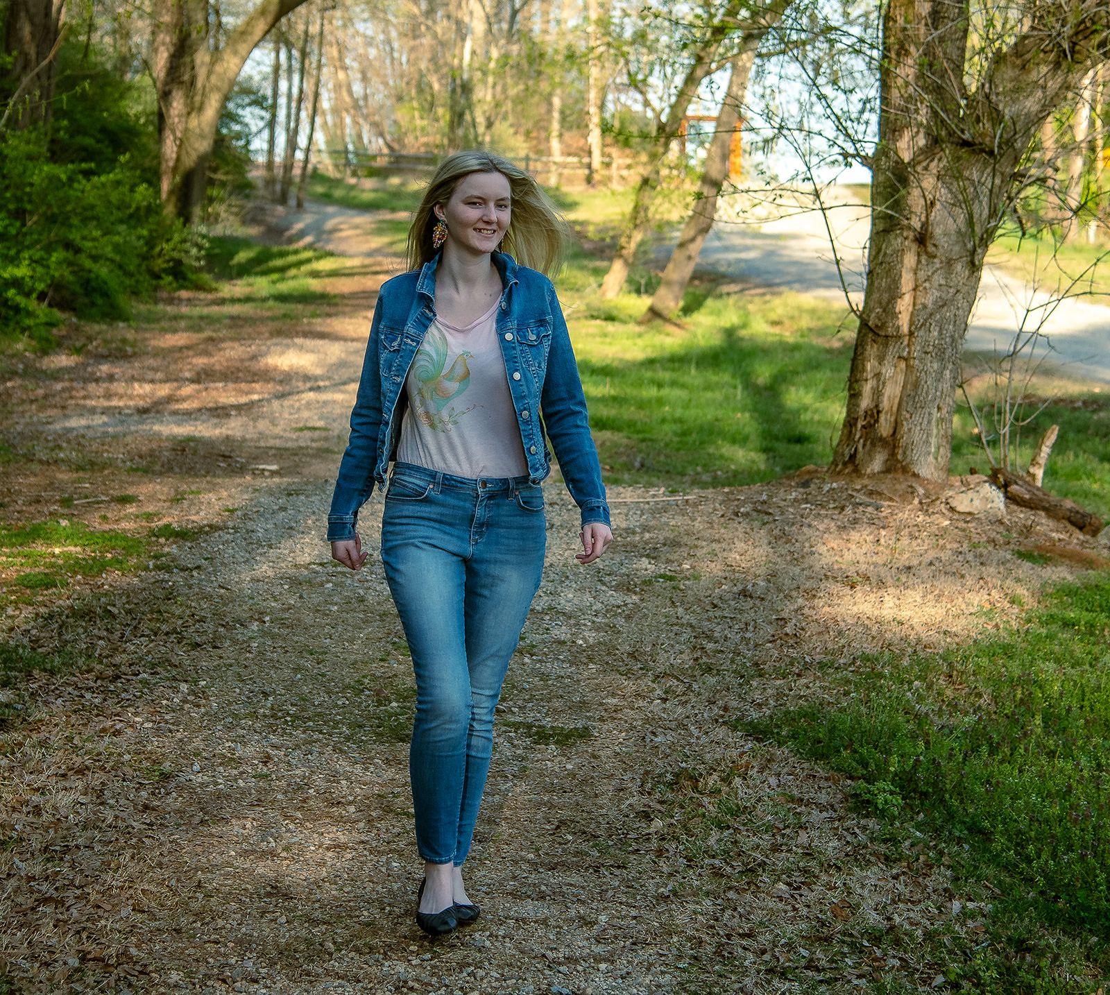 Person walking casually on nature trail wearing denim jacket and jeans on sunny day.