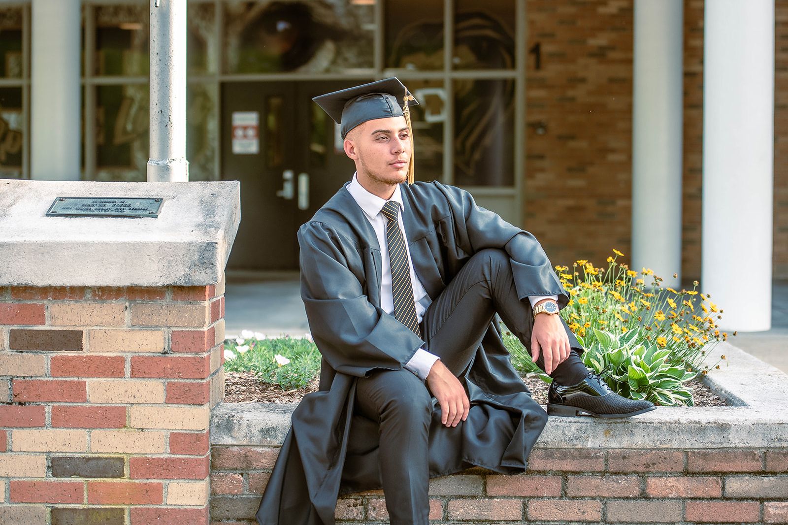 Graduate in traditional black cap and gown sitting on brick wall outside building.