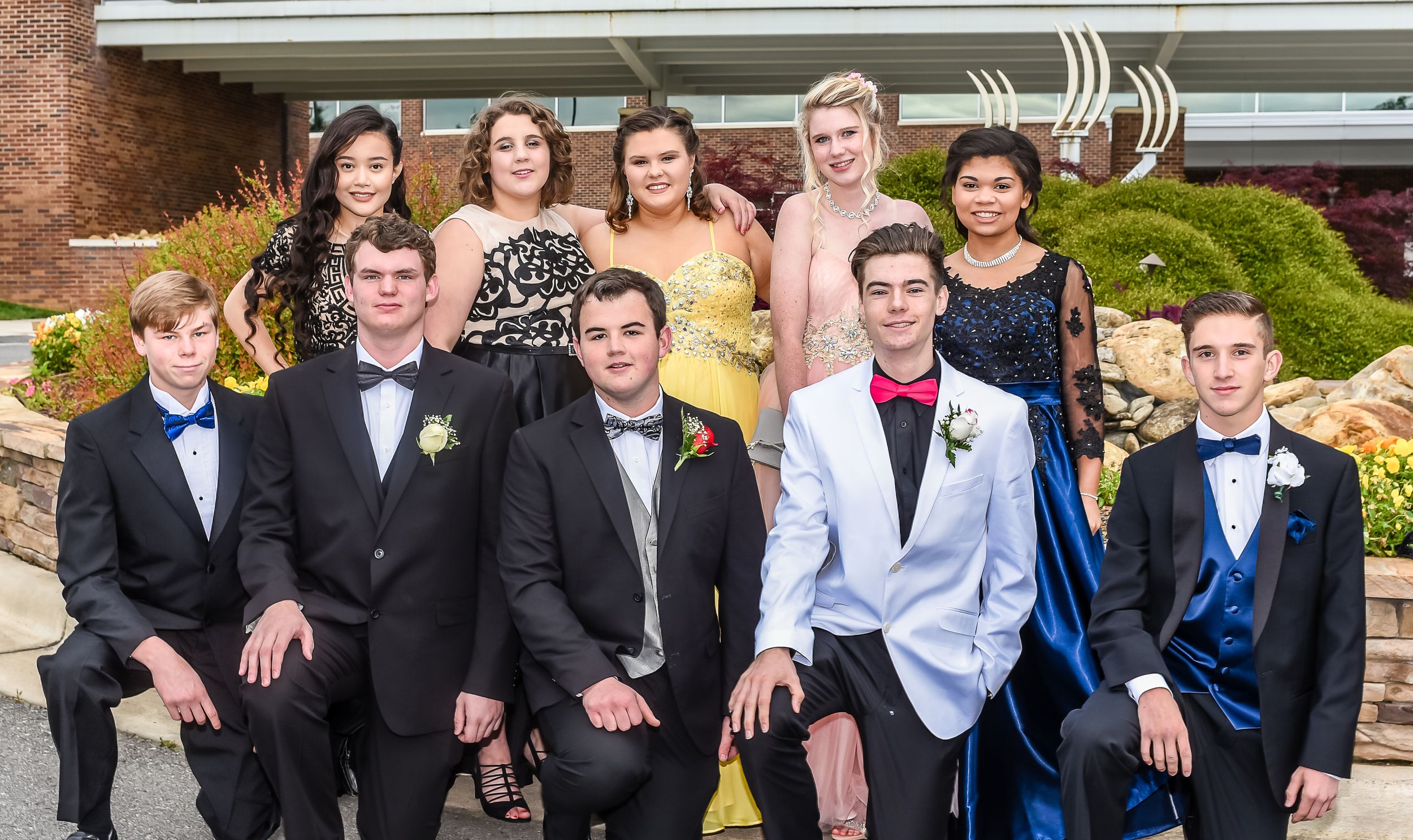 Formal group photo of prom or homecoming attendees in suits and dresses outside brick building.