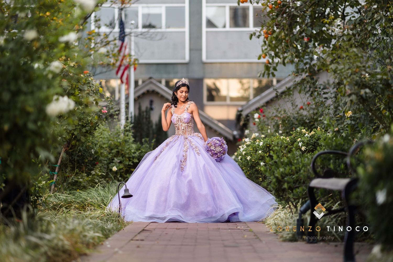 Elegant pose in flowing purple ballgown on garden walkway with American flag in background.