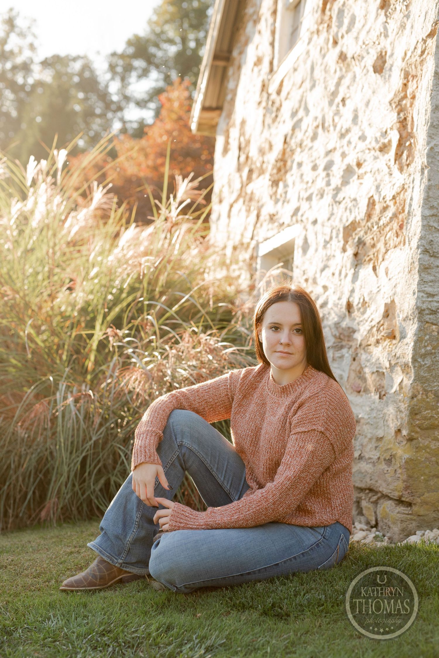 Person in chunky sweater sits casually by rustic stone wall during sunset.