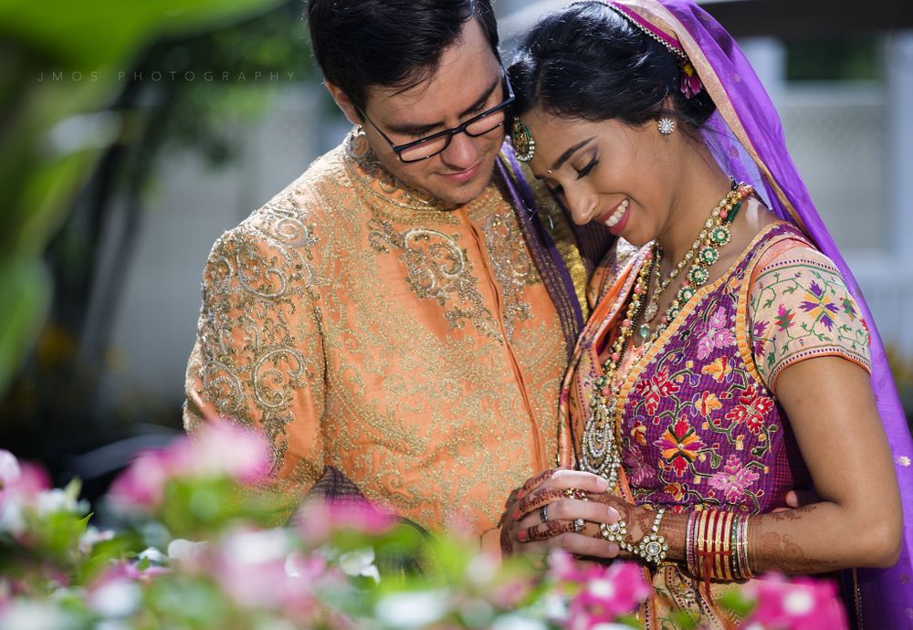 A couple in colorful traditional Indian wedding attire share an intimate moment among pink flowers.