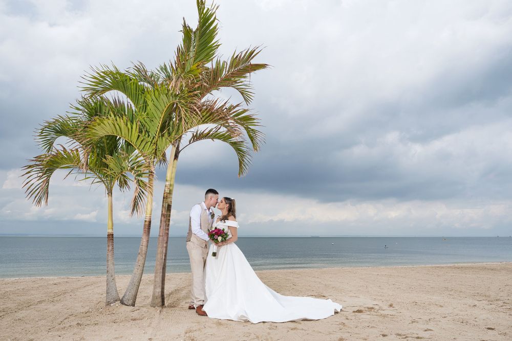 Couple poses by palm trees on a beach with stormy clouds overhead.
