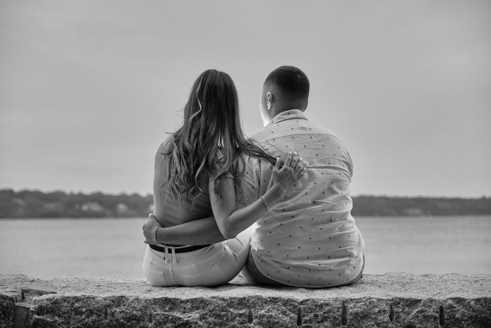 A couple sits together on a stone wall overlooking a serene lake in black and white.