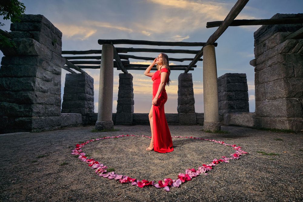 Red dress figure stands in stone structure with rose petals arranged in heart shape.