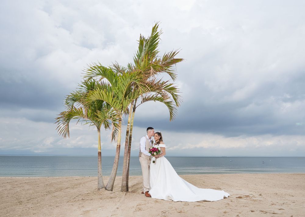 Wedding couple embraces under palm trees on a sandy beach with dramatic sky.