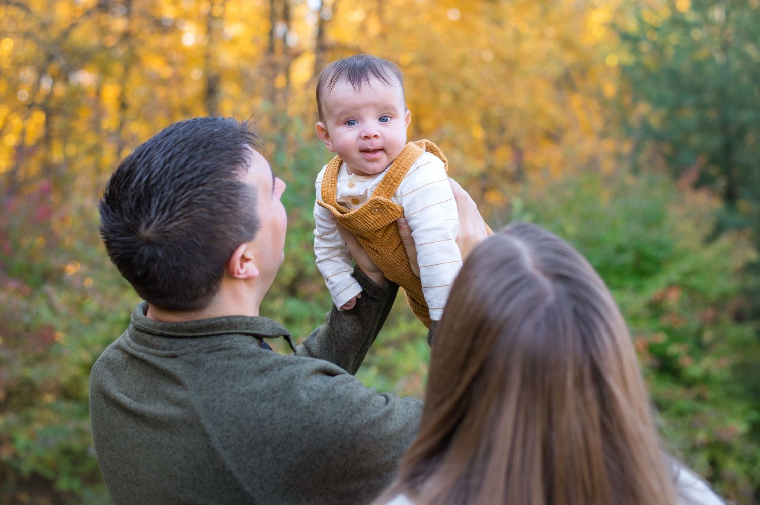 A joyful baby is lifted high by parents during an outdoor autumn photoshoot.
