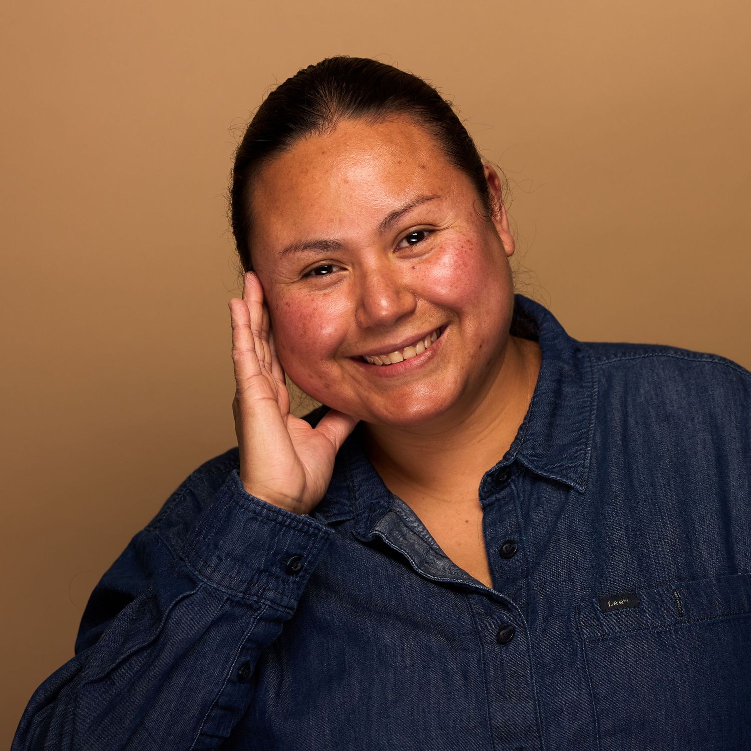Professional headshot of person in denim shirt with hand posed near face, smiling warmly.