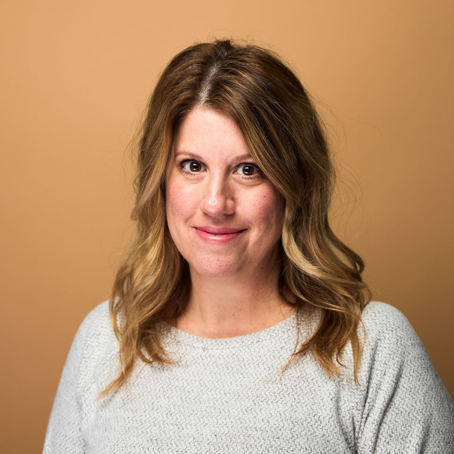 Professional headshot of person with wavy blonde hair wearing light gray sweater against brown background.