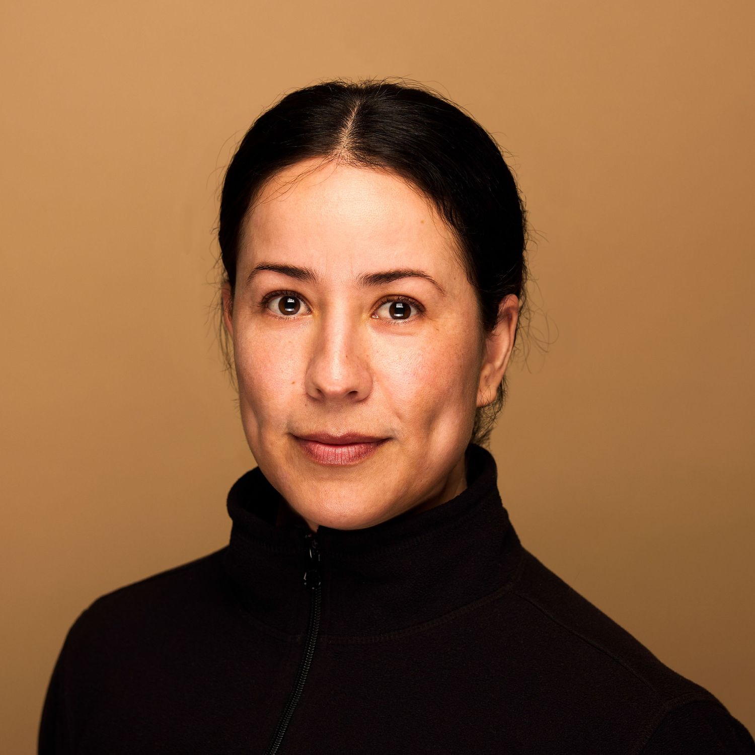 Portrait of person in black turtleneck with hair pulled back against brown background.