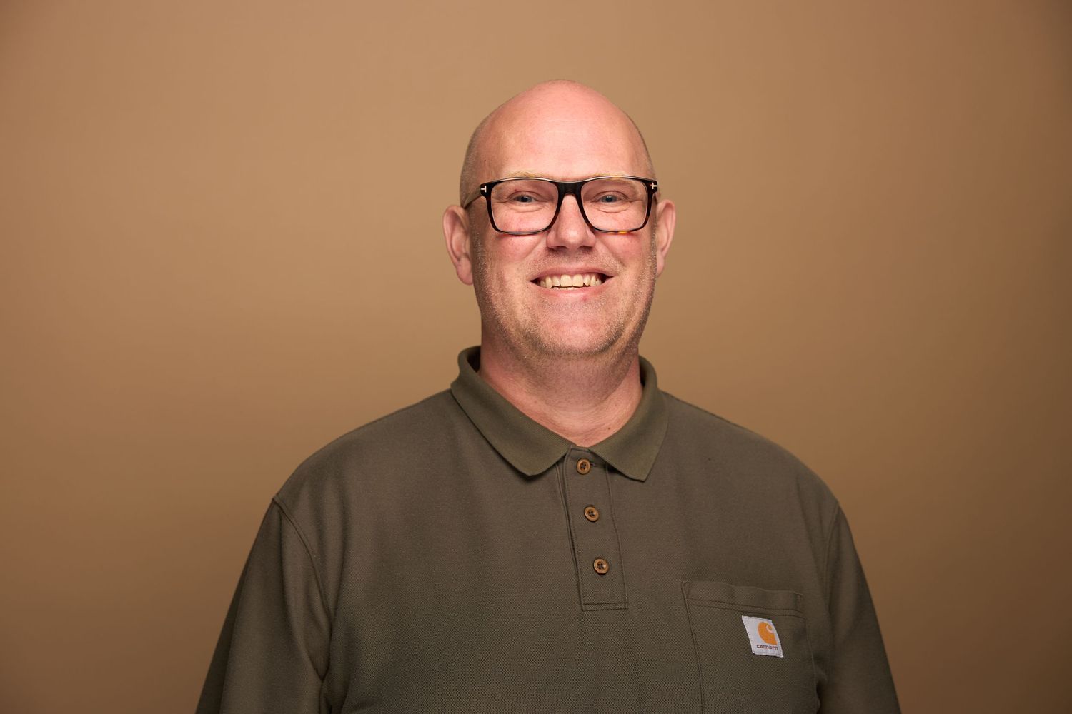 Portrait of a bald man wearing glasses and a brown Carhartt polo shirt against brown background.
