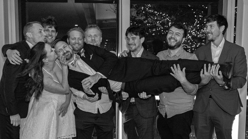 Black and white photo of wedding party playfully supporting the bride on outdoor deck.