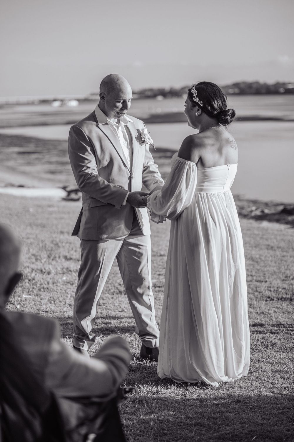 A monochrome wedding photo shows a couple holding hands during their ceremony by the water.