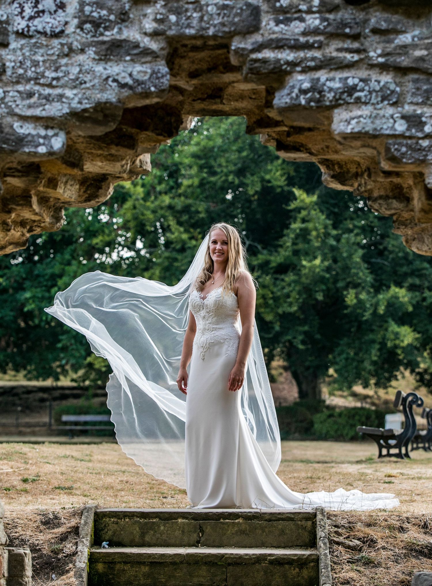 A flowing white wedding dress billows dramatically against a stone wall backdrop in an outdoor setting.