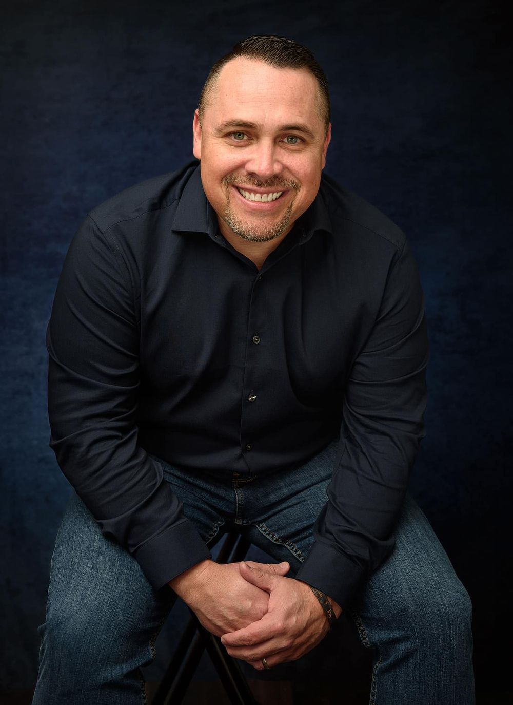 Professional portrait of man in black button-down shirt against dark background.