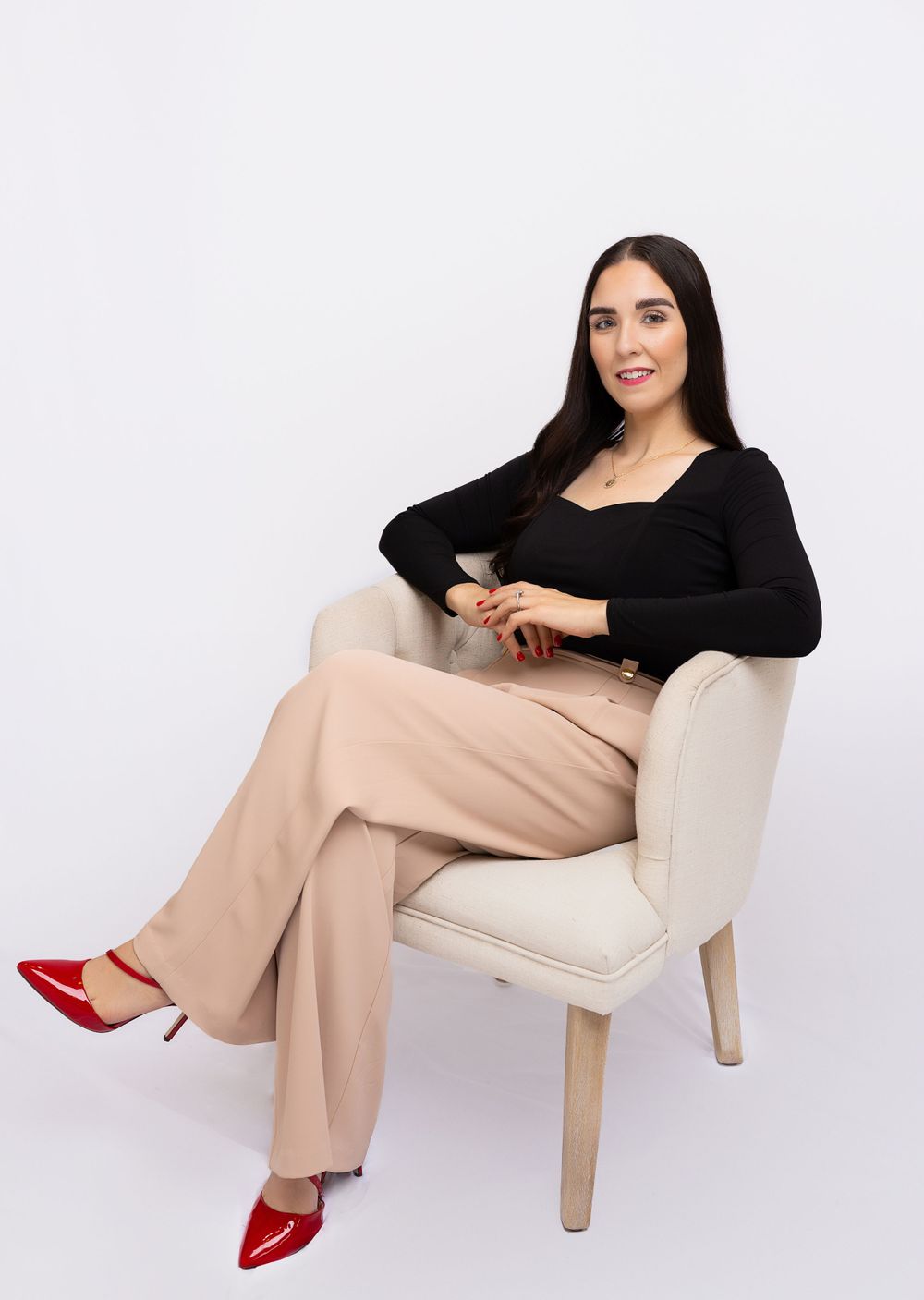Person in black top and beige pants sitting in cream colored chair wearing red shoes.