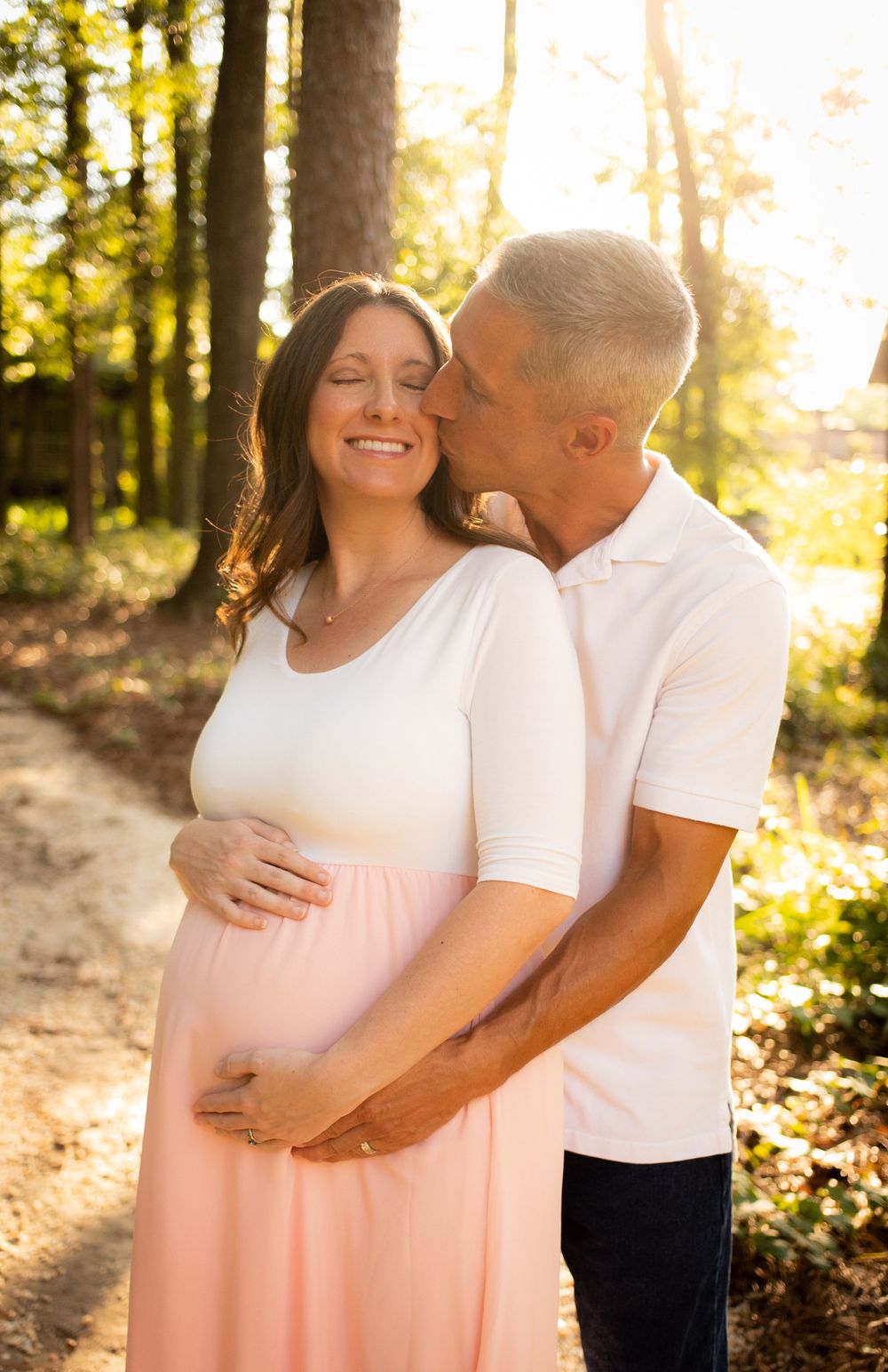 Pregnant woman smiling as man kisses her cheek in a sunlit forest path, hands on belly.