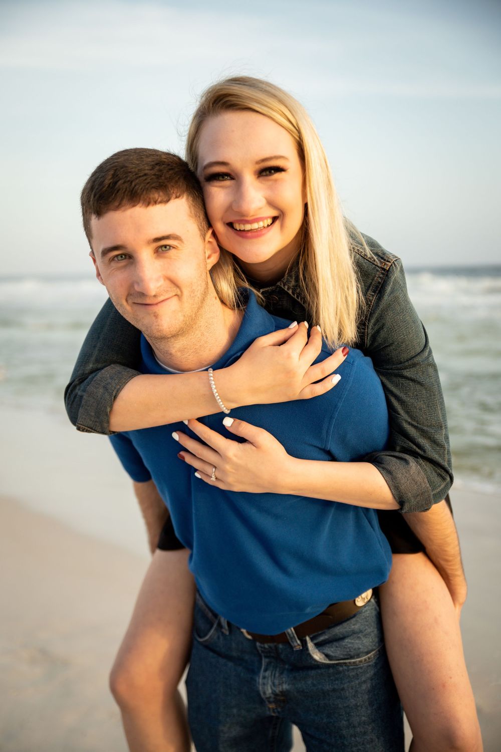 Happy couple posing on the beach, woman giving piggyback ride smiling at camera with ocean waves in background.